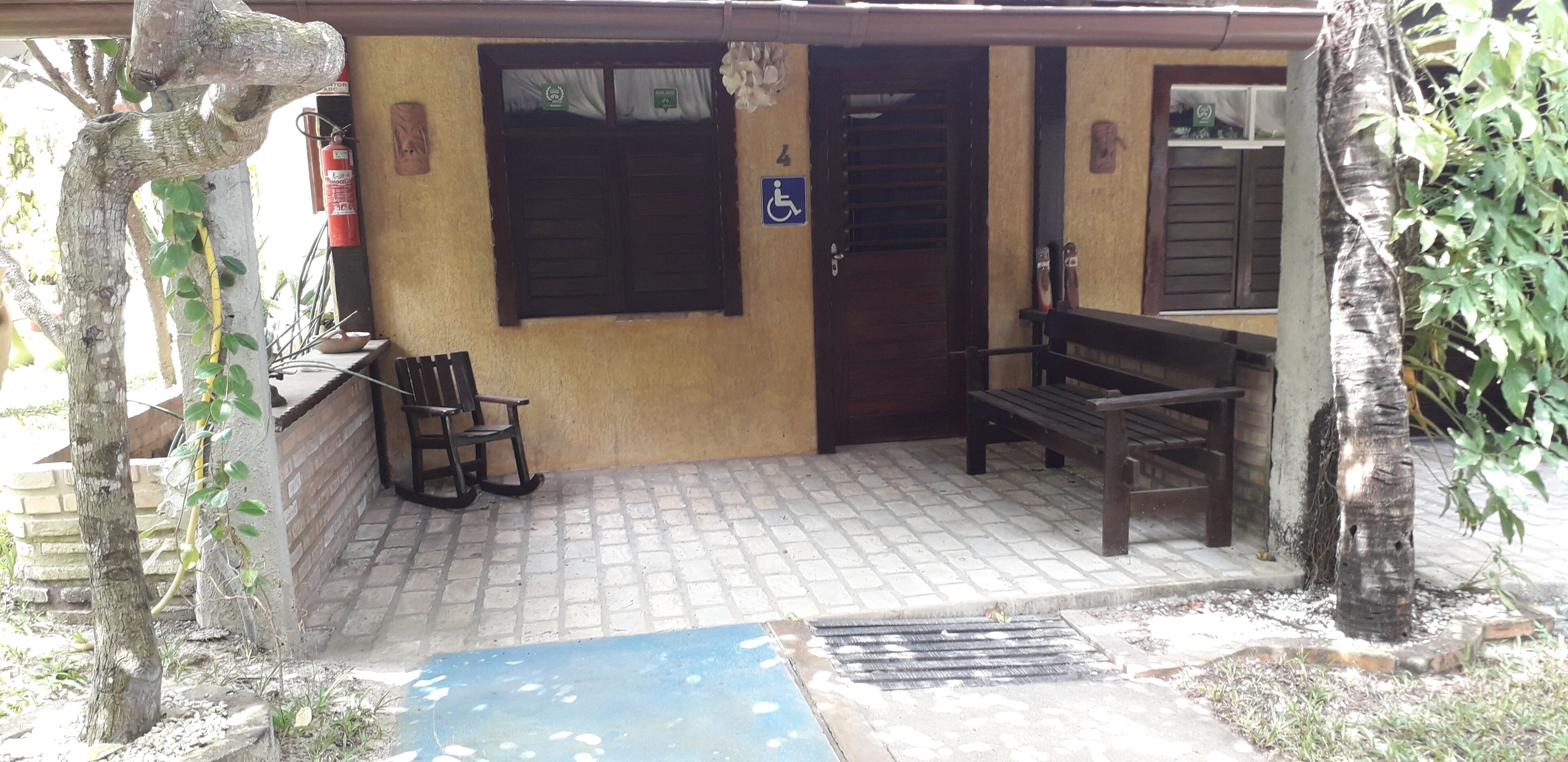 Un espacio de estar al aire libre con techo, que incluye un banco y sillas, una puerta con señal de accesibilidad para sillas de ruedas y un ventanal con cortina.