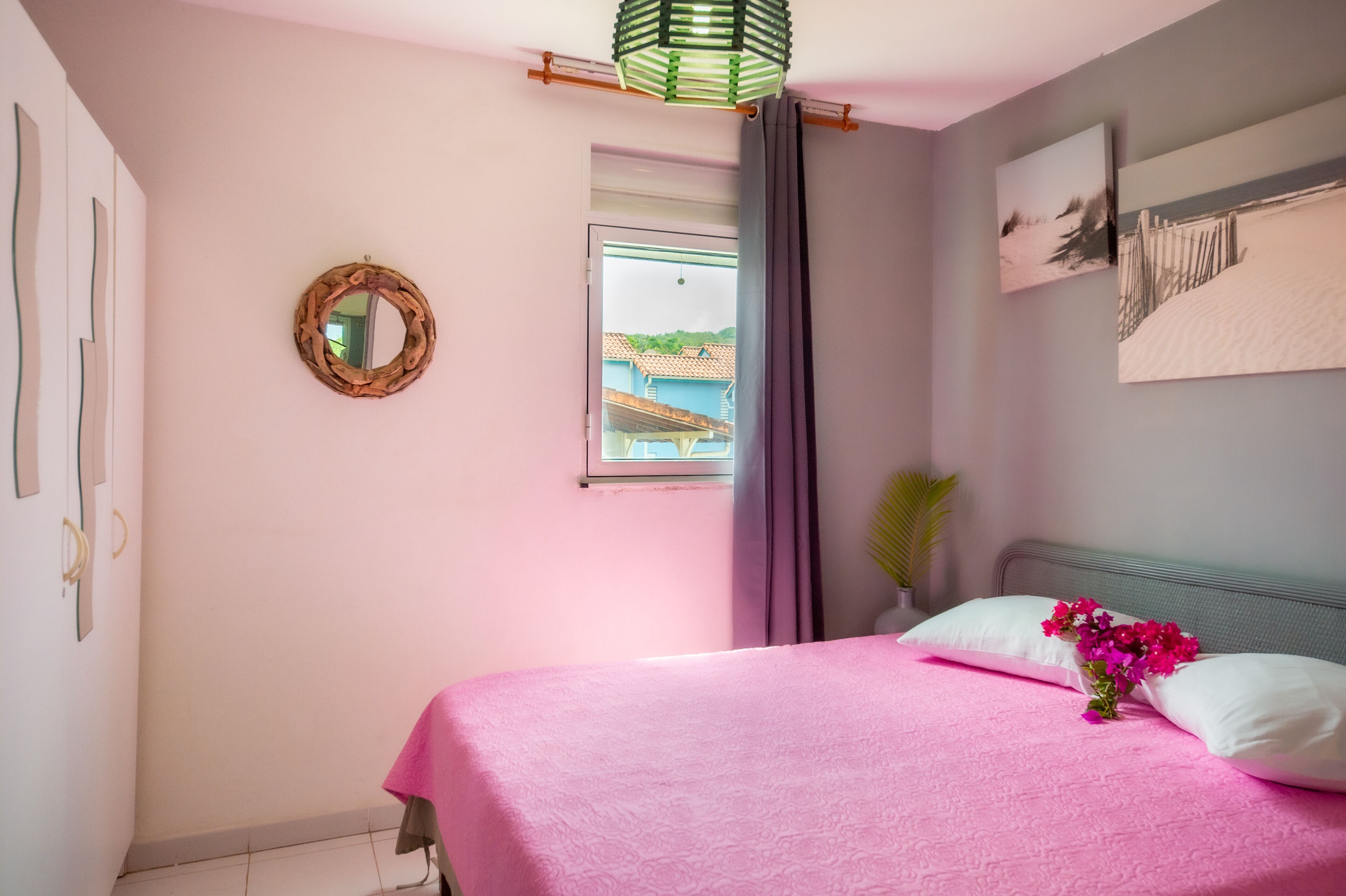 A bedroom with a pink bedspread, a mirror, a window with curtains, and a wall-mounted picture.