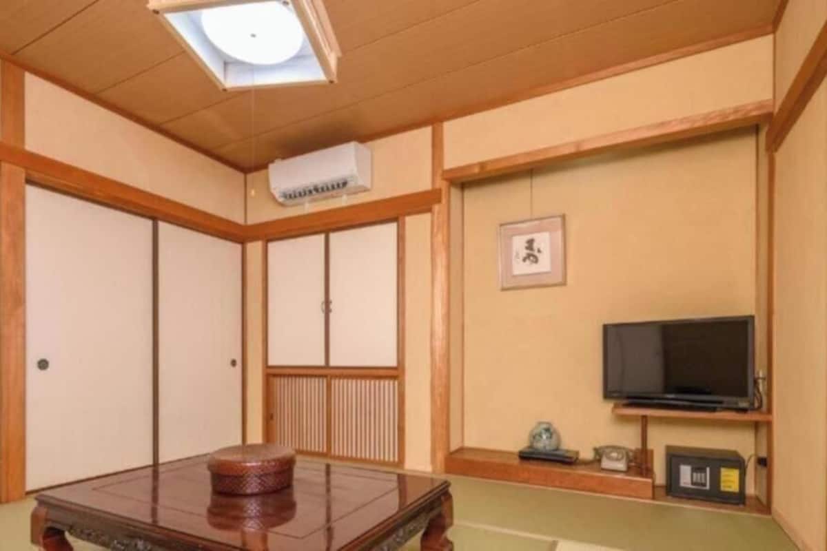Una habitación japonesa tradicional con suelo de tatami, una mesa de madera baja, puertas correderas, un televisor y un estante con objetos.