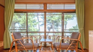 Una habitación con dos sillones de mimbre y una mesita, ventanas amplias con cortinas verdes y vistas a árboles.