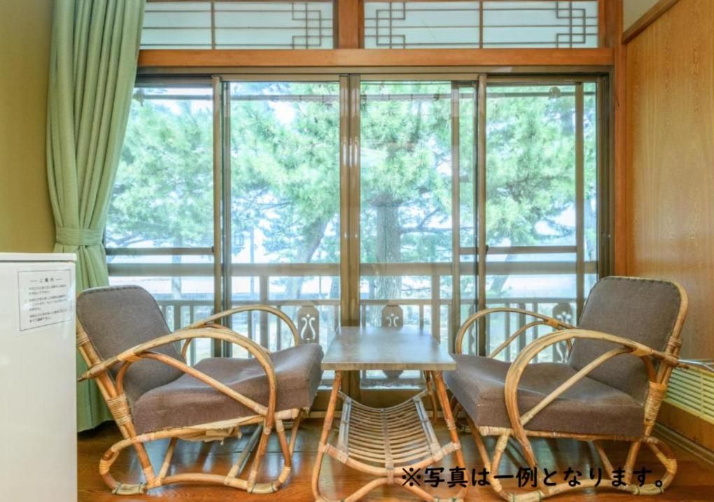 A room with a wooden table and two wicker chairs, a window with a view of trees, and a green curtain.