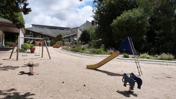 Un'area giochi con altalene e scivolo, circondata da alberi e un edificio sullo sfondo.