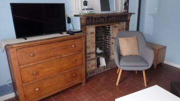 Uma sala de estar com uma TV sobre um aparador de madeira, uma lareira com uma estatueta de pássaro, uma poltrona cinza e uma pequena mesa de madeira.