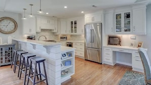 A modern kitchen with white cabinetry, a stainless steel refrigerator, and a bar area with stools. - 2 Blocks from Beach, Dog Friendly w/ no dog fees!, Hot Tub, Fenced Yard (Kure Beach)