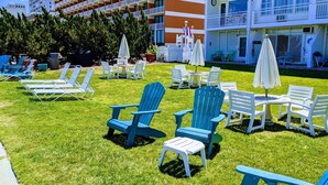 Une cour d’hôtel avec des espaces de détente en plein air, comprenant des chaises Adirondack bleues et des tables blanches, le tout entouré d’une pelouse bien entretenue et d’un immeuble à plusieurs étages doté de balcons.