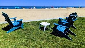 Un espace verdoyant avec deux chaises Adirondack bleues et une petite table blanche, d’où l’on peut admirer une plage avec des parasols et des personnes au loin.