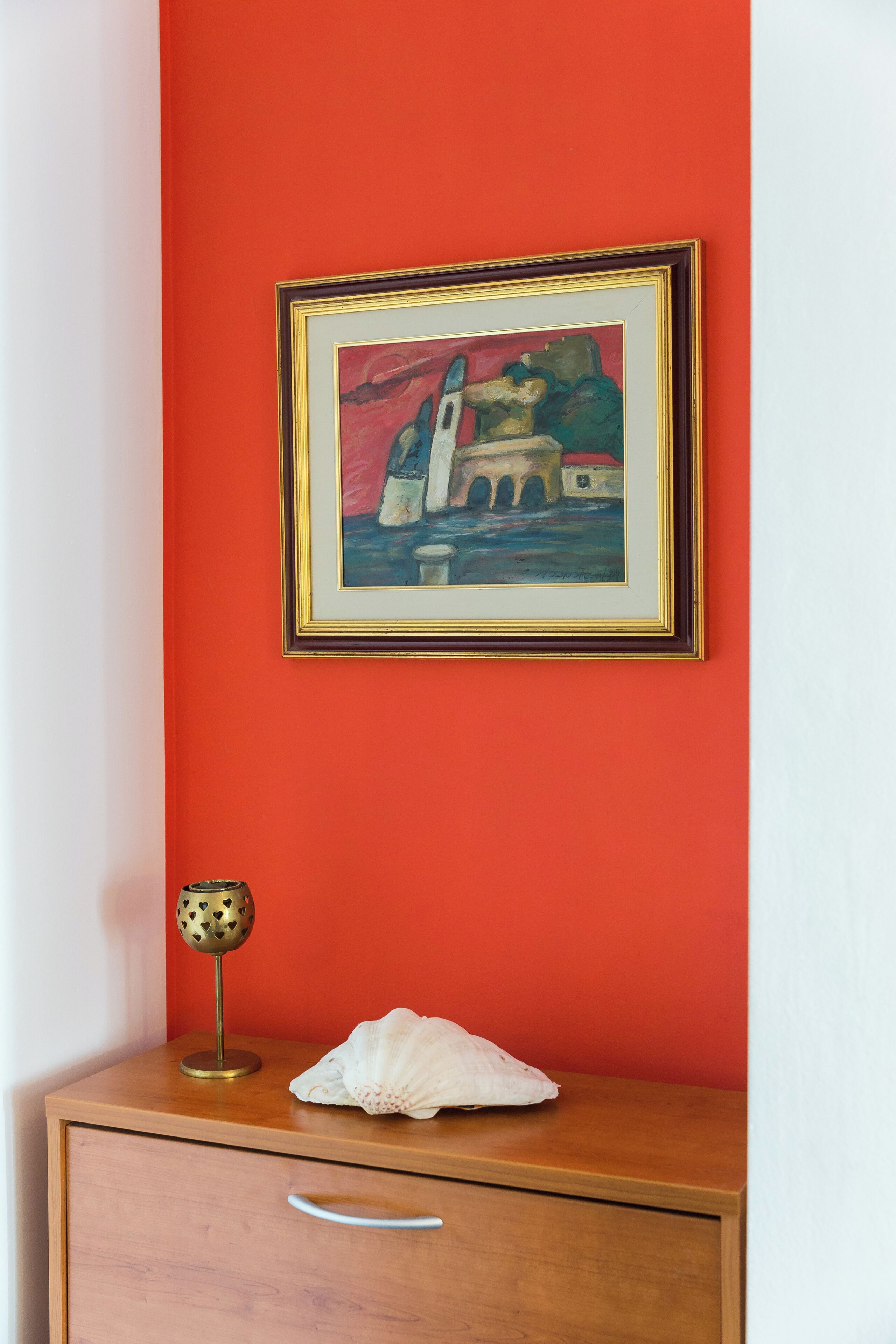 A framed painting of a building on a red wall, a brass lamp, and a white object on a wooden dresser.