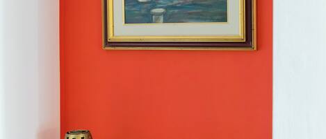 A framed painting of a building on a red wall, a brass lamp, and a white object on a wooden dresser.