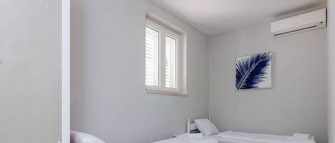 Ein kleines, sauberes Zimmer mit zwei weißen Betten, einem Fenster und einer Wand-Klimaanlage.