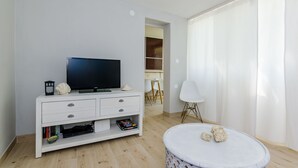 A modern living room with a flat-screen TV on a white TV stand, a white armchair, and a round coffee table with decorative items. - Ploce Apartments - Kate (Dubrovnik)