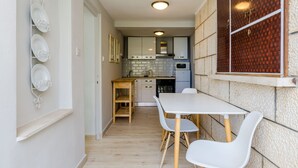 A compact kitchen with a dining area, featuring a white table and chairs, a wooden shelf, and a tiled backsplash. - Ploce Apartments - Kate (Dubrovnik)