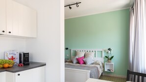Een compacte kamer met kitchenette, een bed met wit hoofdbord, een klein nachtkastje met een plant, en een ontbijtgedeekte eettafel.