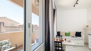 Een balkon met een stoel en een tafel, een bed met kussens, en een kleine kitchenette.