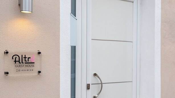 Een witte deur met een ruit, een wandlamp en een bordje met 'Altr Guest House' en contactgegevens.