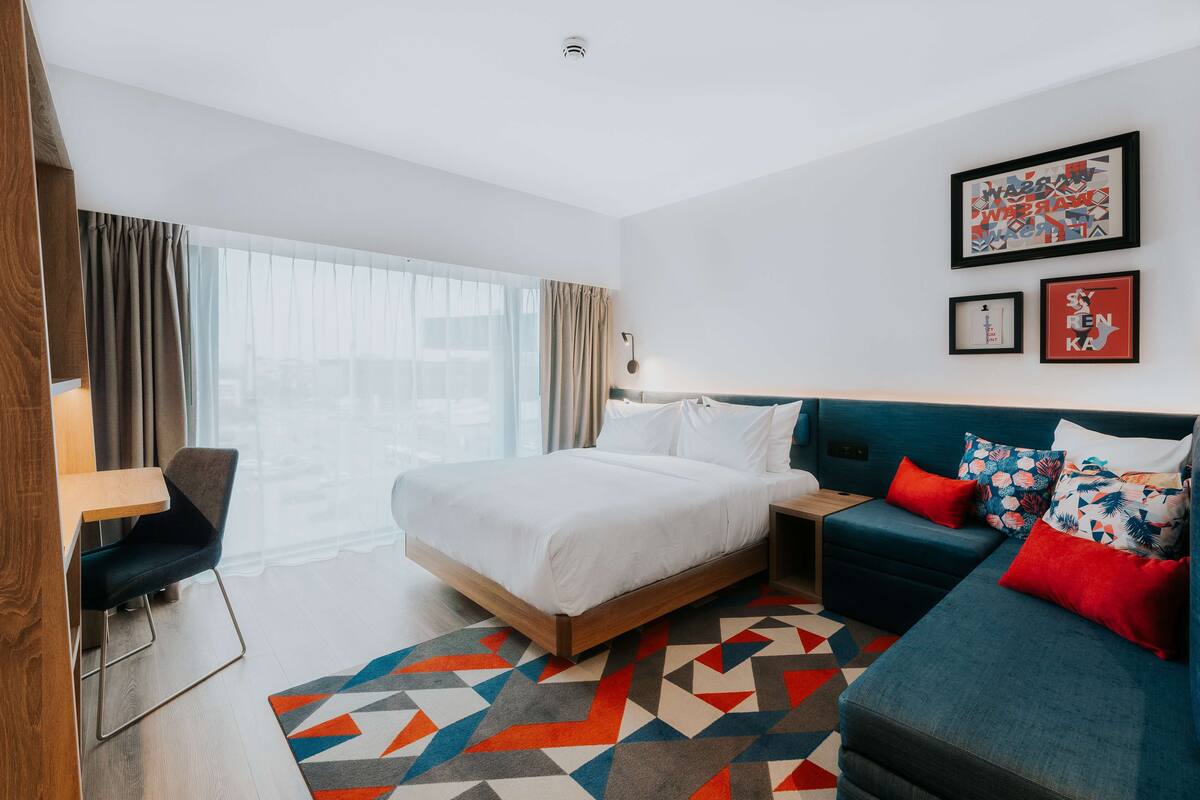 A modern hotel room with a large bed, a sofa, a desk, and a view of the city.