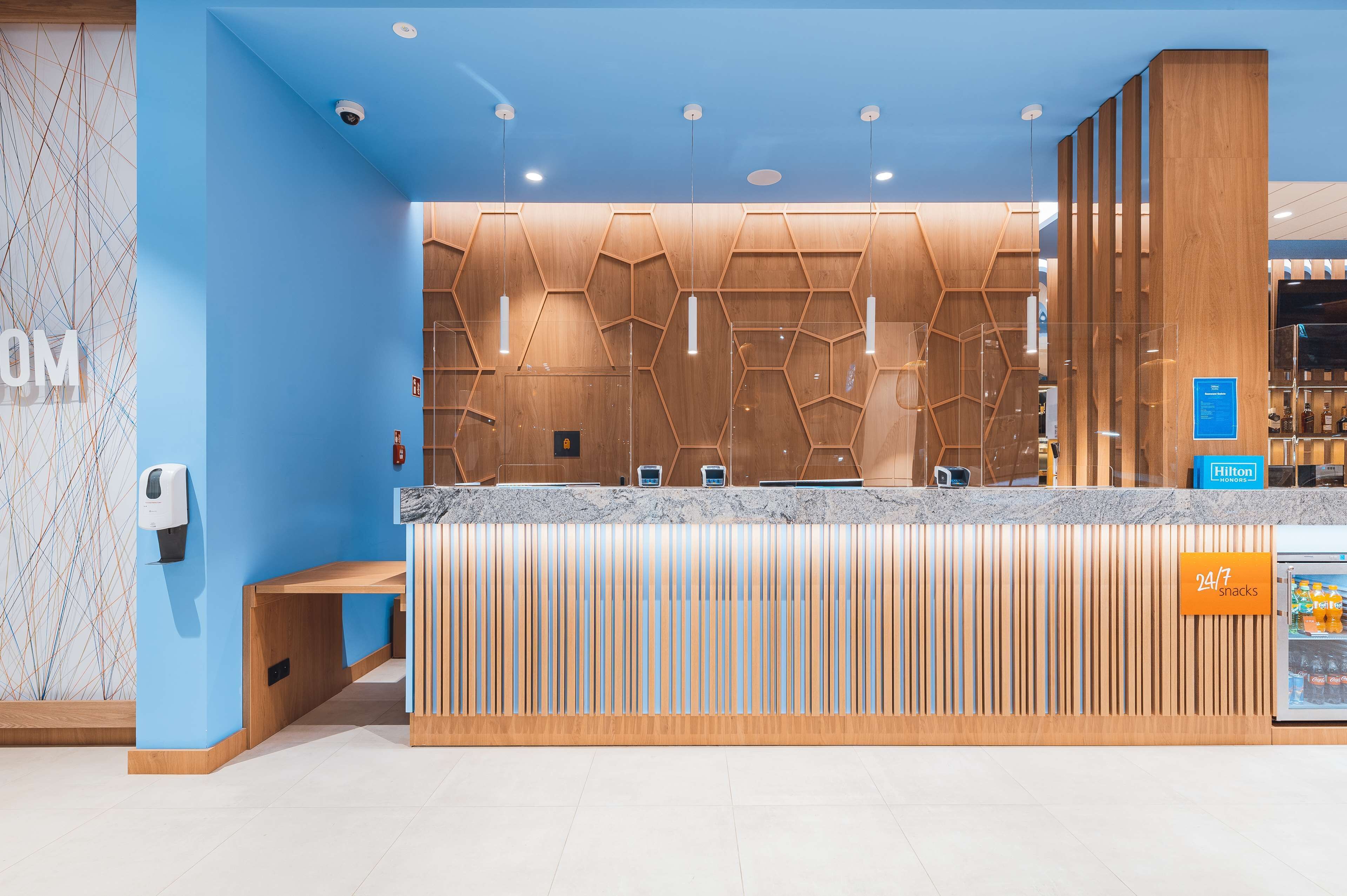 A hotel reception area with a modern design, including a reception desk, a 24/7 snack bar, and a security camera.