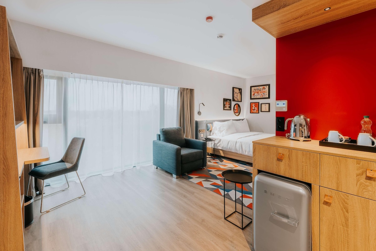 Una habitación de hotel moderna con cama, escritorio, silla, kitchenette y un ventanal con cortinas.