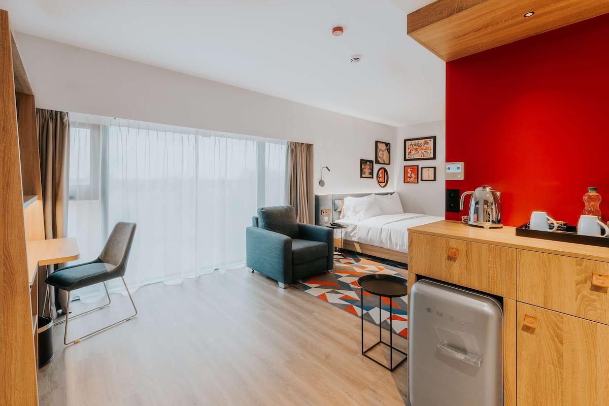 A modern hotel room with a bed, a desk, a chair, a kitchenette, and a large window with curtains.