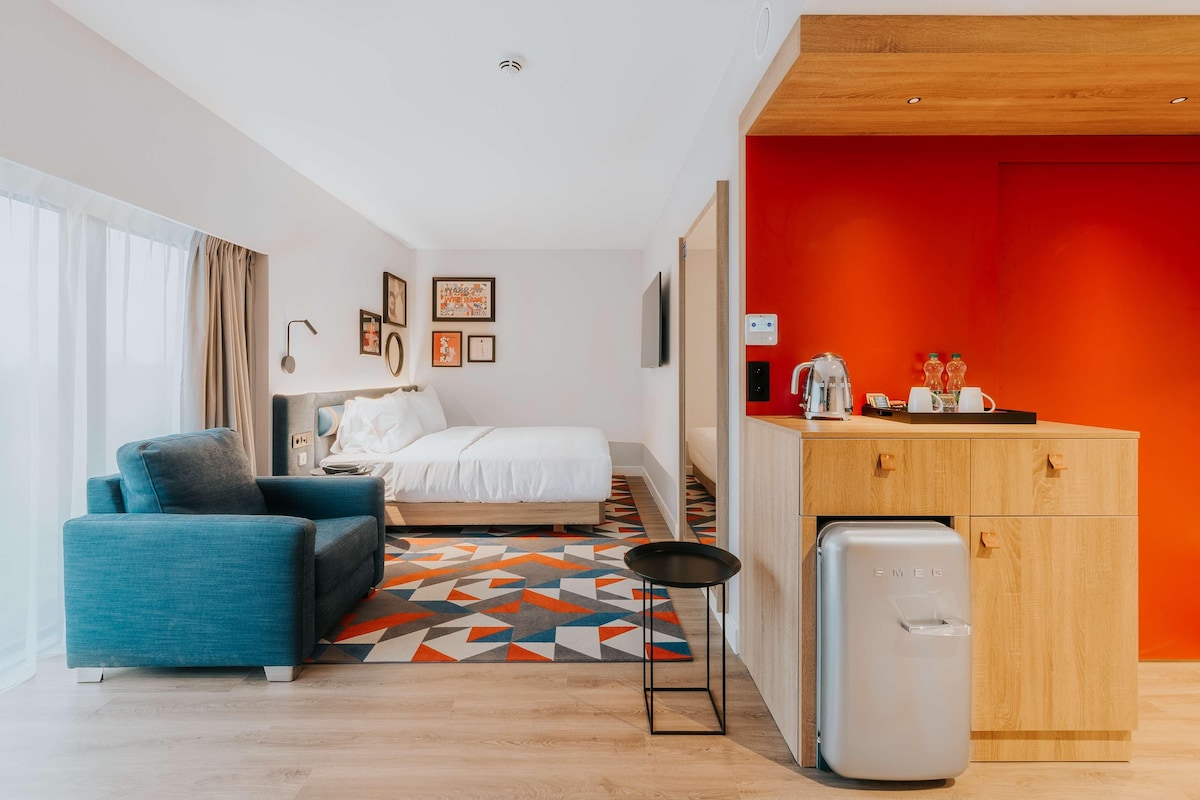 A modern hotel room with a bed, a blue sofa, a small kitchen area with a fridge, and a geometric patterned rug.