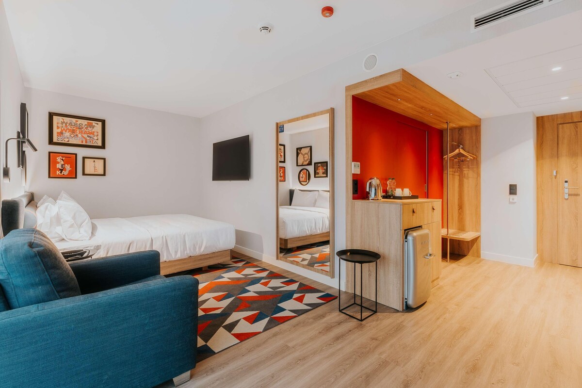 A modern hotel room with a bed, a blue armchair, a small kitchen area, and a mirror reflecting the room.