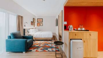 A modern hotel room with a bed, a blue sofa, a small table, and a kitchenette with a refrigerator.