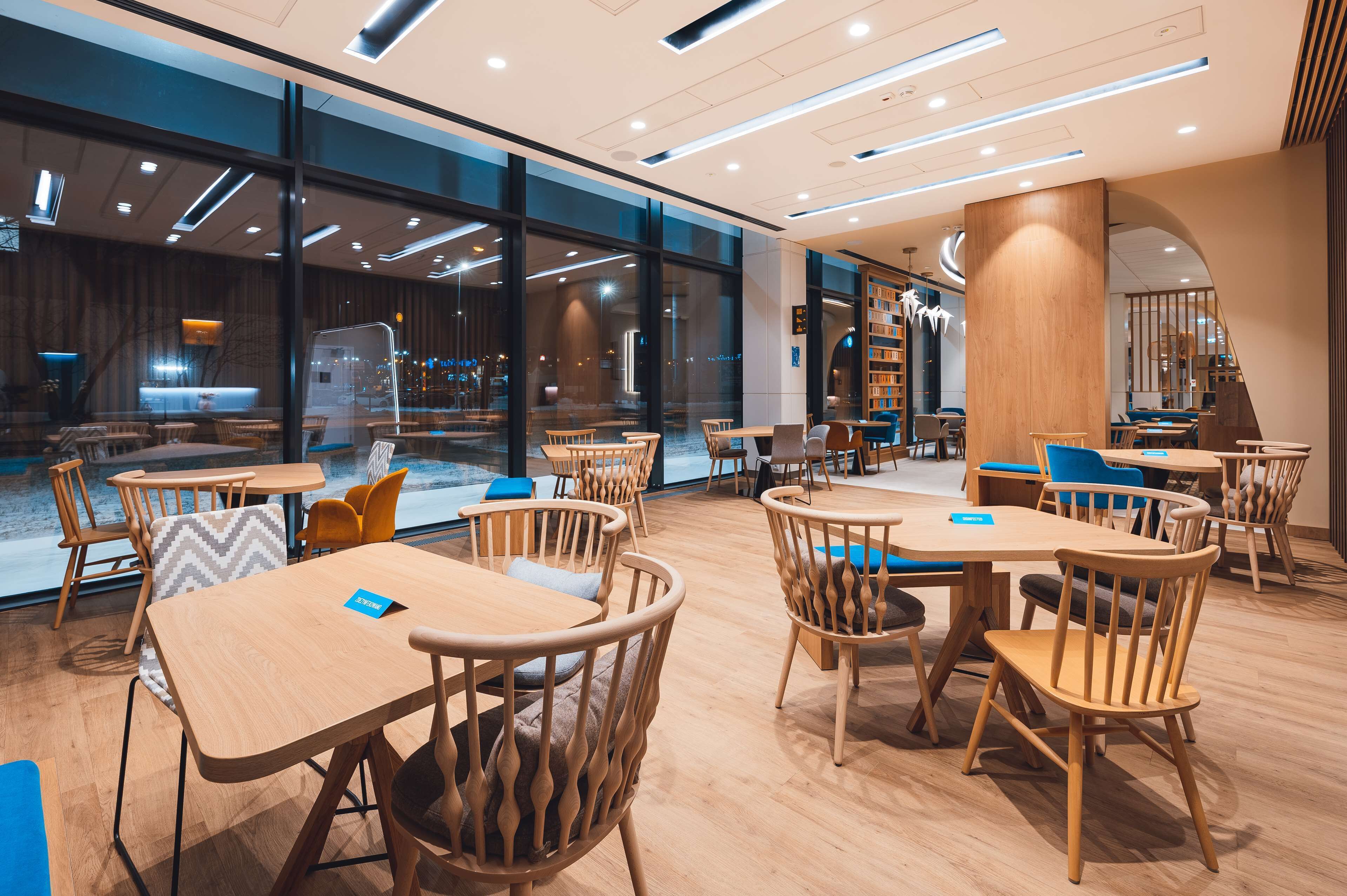 A modern indoor dining area with wooden tables and chairs, large windows, and a visible staircase.