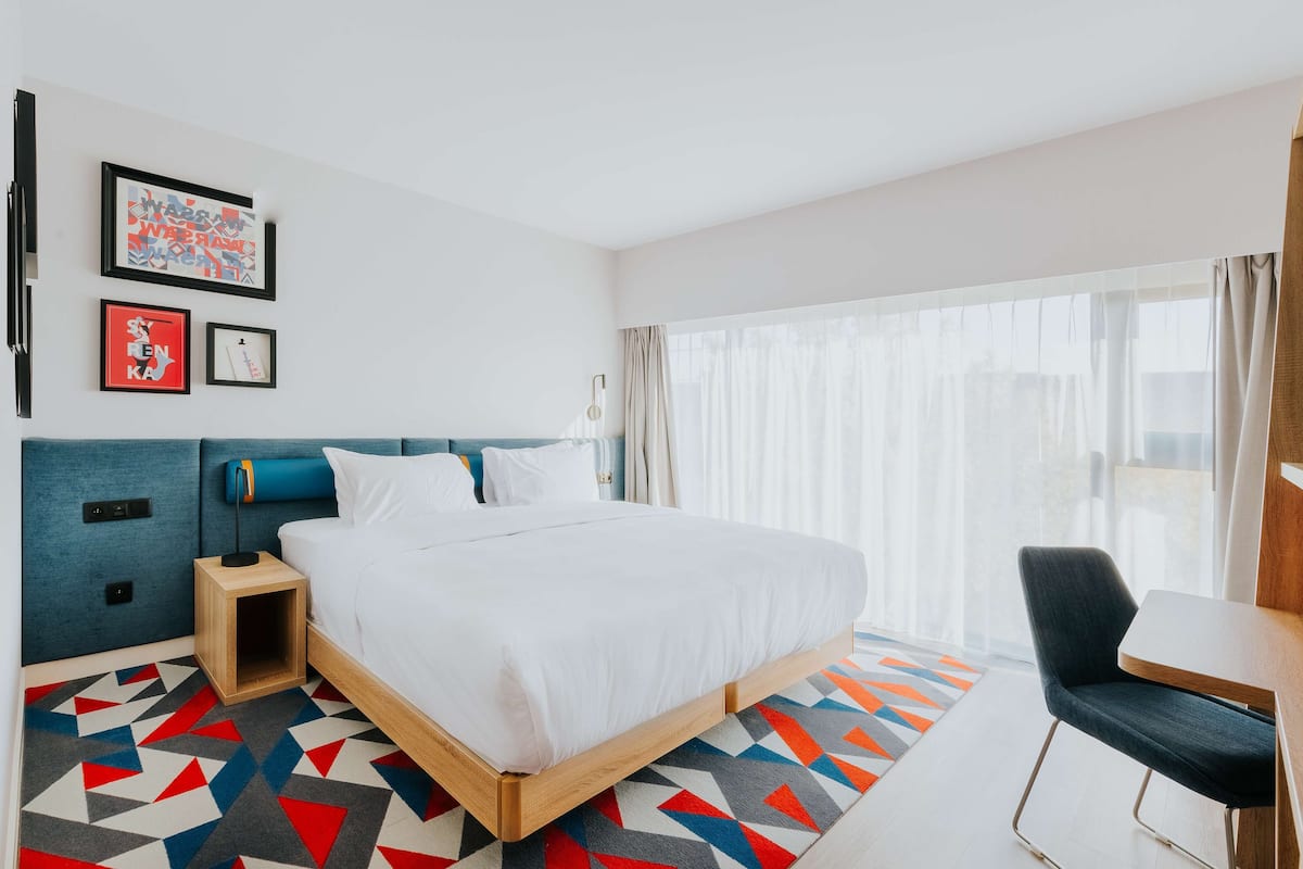 A modern hotel room with a large bed, a desk, and a chair. There are framed pictures on the wall, a nightstand, and a window with curtains.