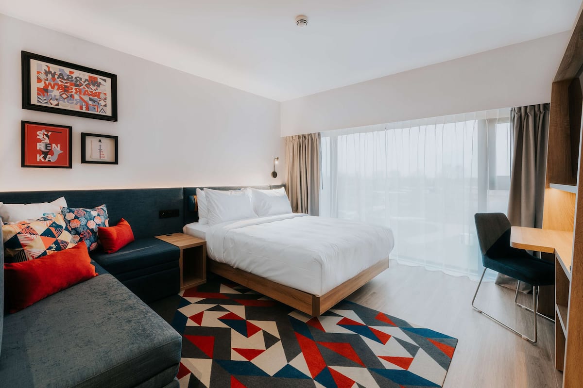 A modern hotel room with a large bed, a sofa, a desk, and a view of the city through large windows.