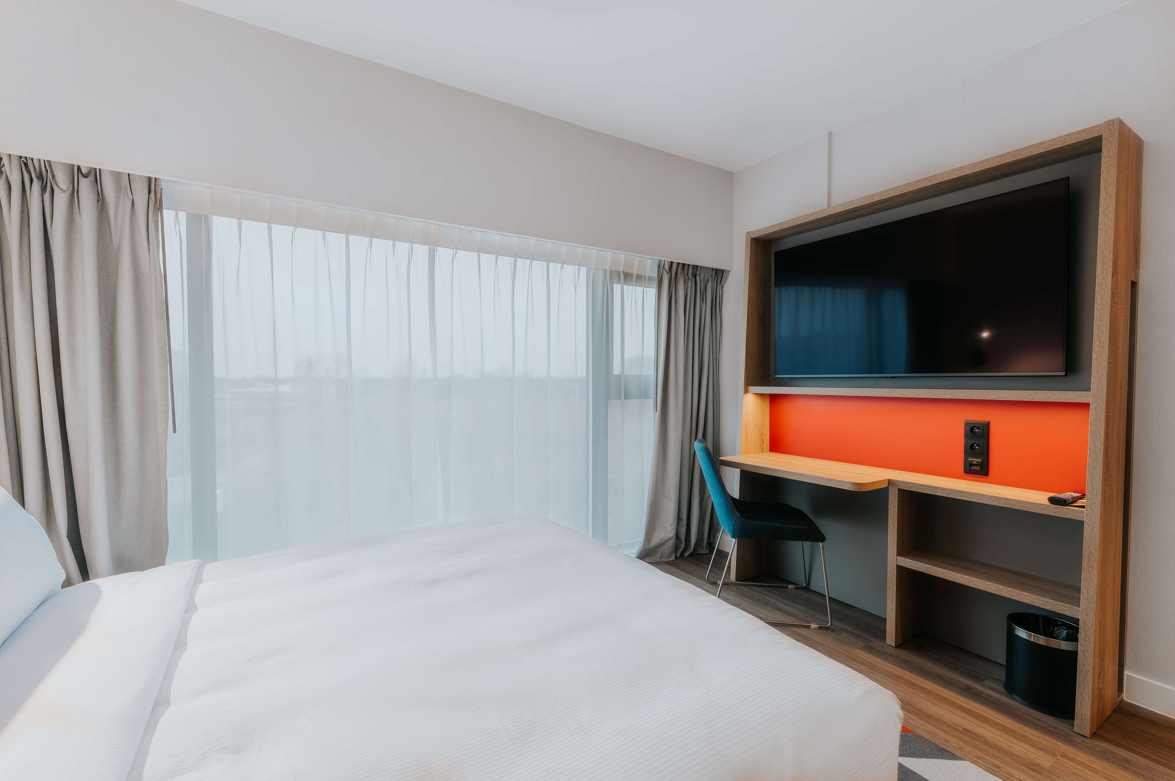 Una habitación de hotel con cama, escritorio con televisor y vista a la ciudad a través de amplios ventanales.
