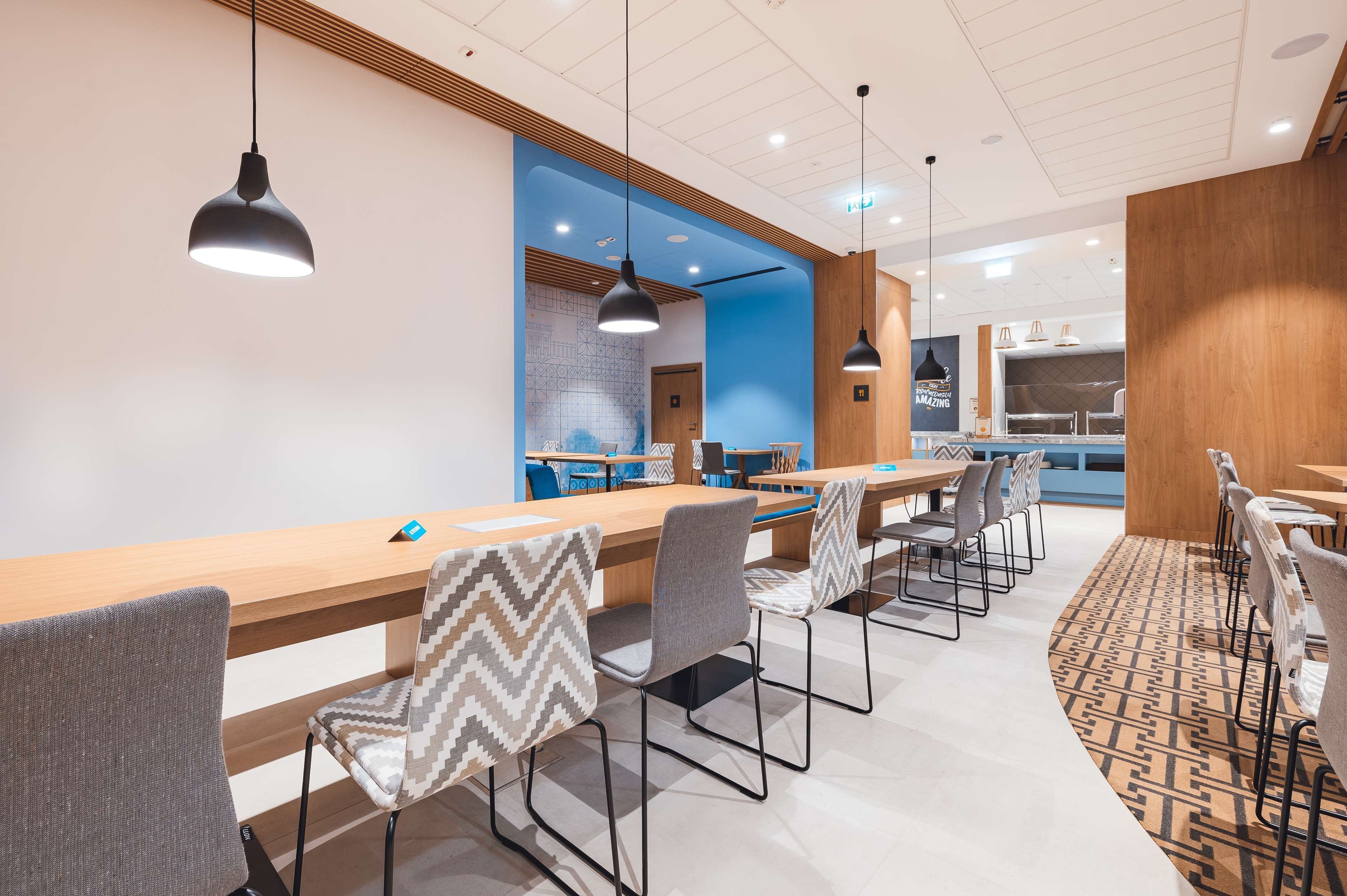 A modern dining area with a long wooden table, grey chairs with patterned cushions, and pendant lighting.