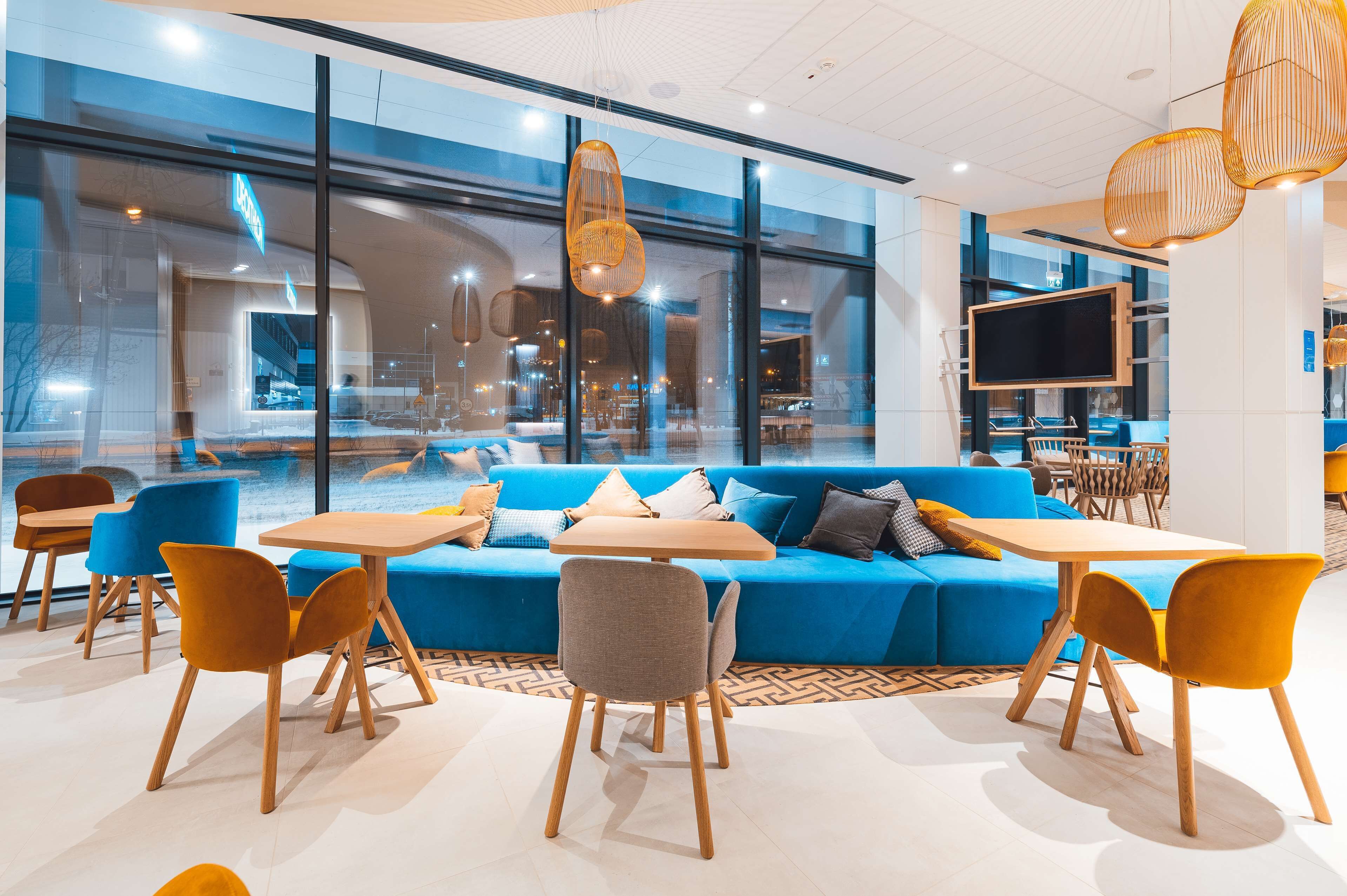 Un interior moderno con un sofá azul, mesas y sillas de madera, ventanas amplias y lámparas colgantes de mimbre.