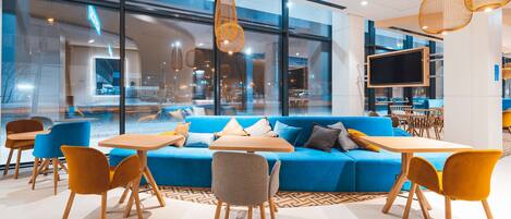 Un interior moderno con un sofá azul, mesas y sillas de madera, ventanas amplias y lámparas colgantes de mimbre.