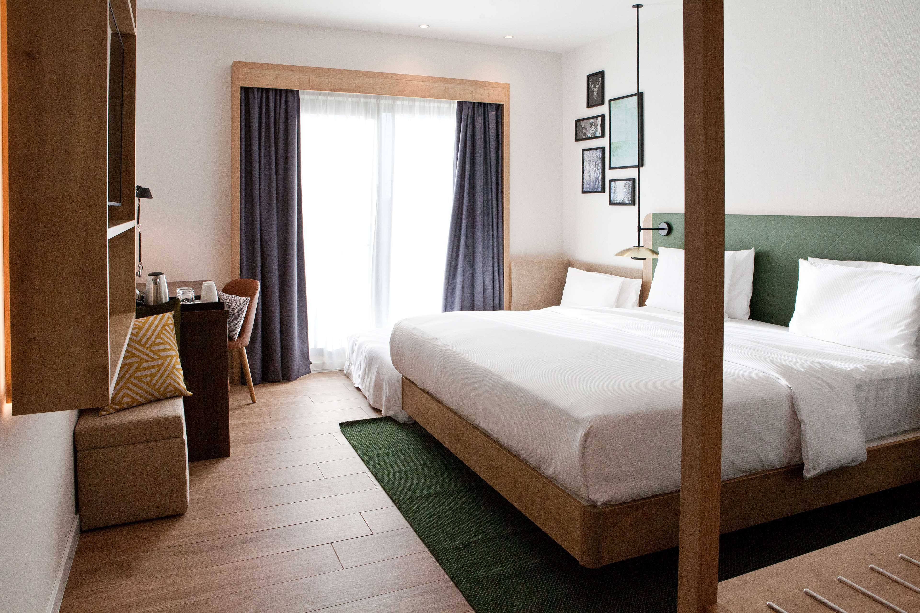 A hotel room with a large bed, a desk with a chair, a small ottoman, and a window with curtains.