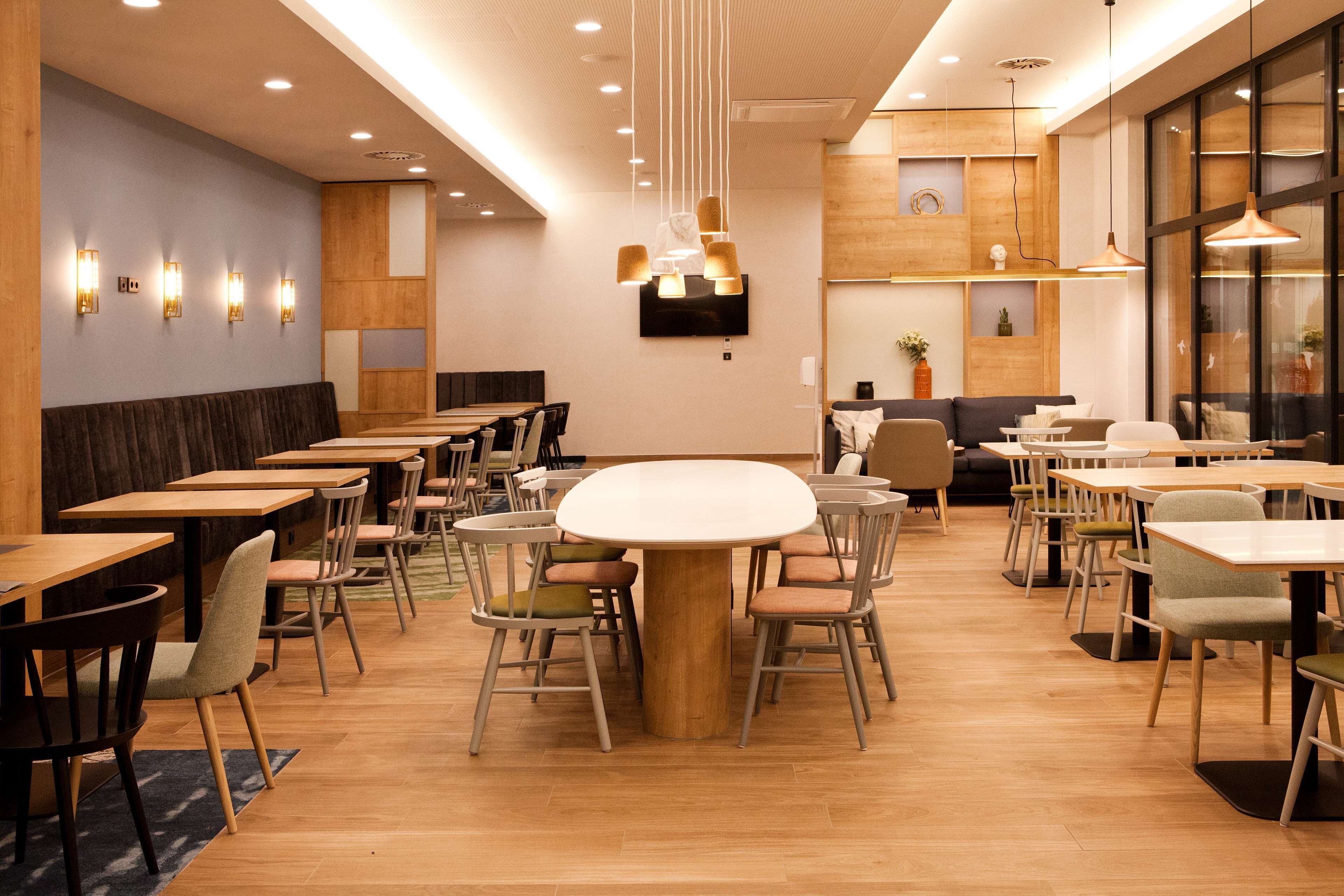 A modern restaurant interior with wooden tables and chairs, a central round table, and pendant lighting.
