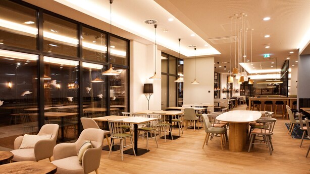 En modern restaurang med träbord och stolar, hängande lampor och stora fönster som erbjuder utsikt över staden på kvällen.