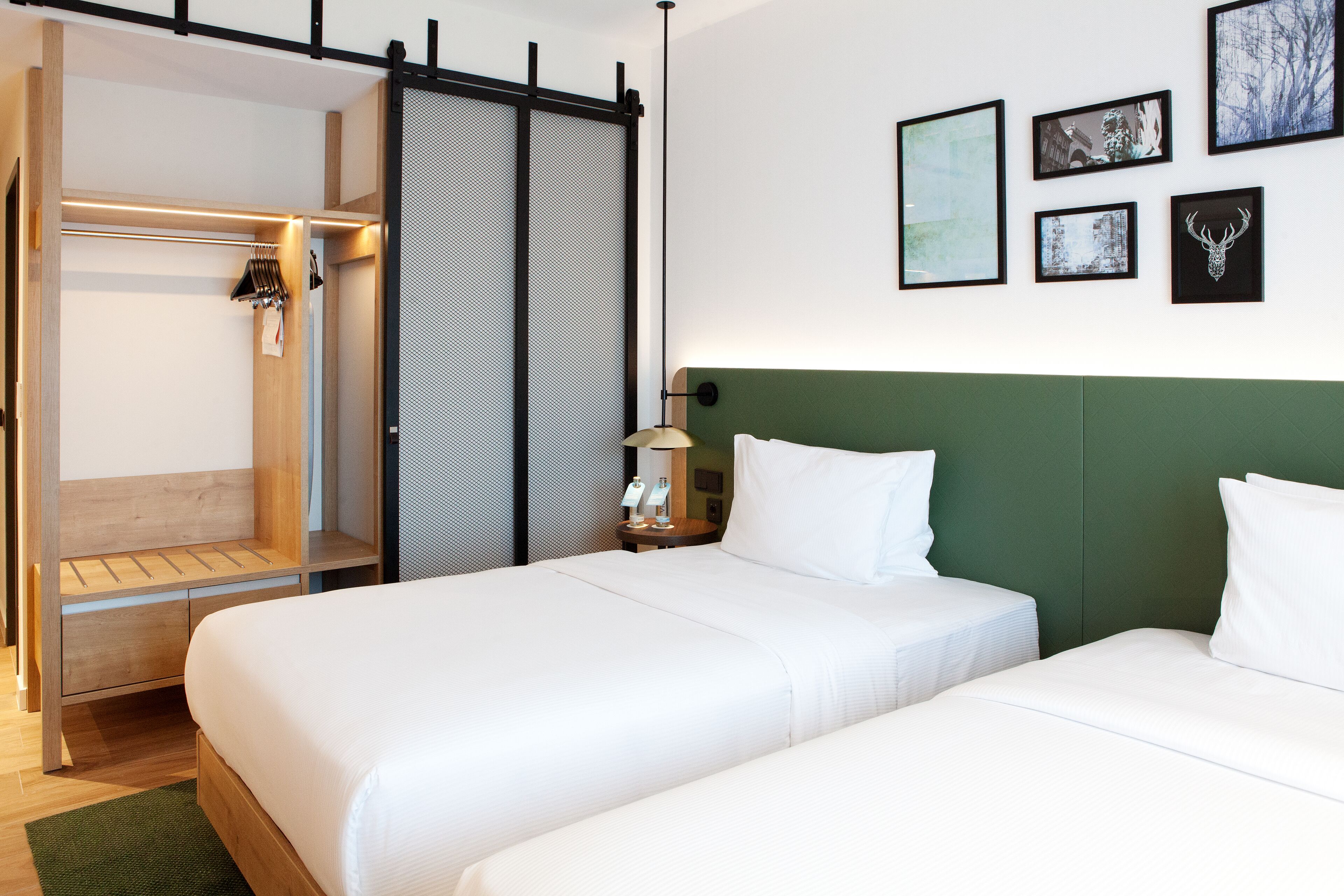 A hotel room with two beds, a wardrobe, a desk, and a wall with framed pictures.