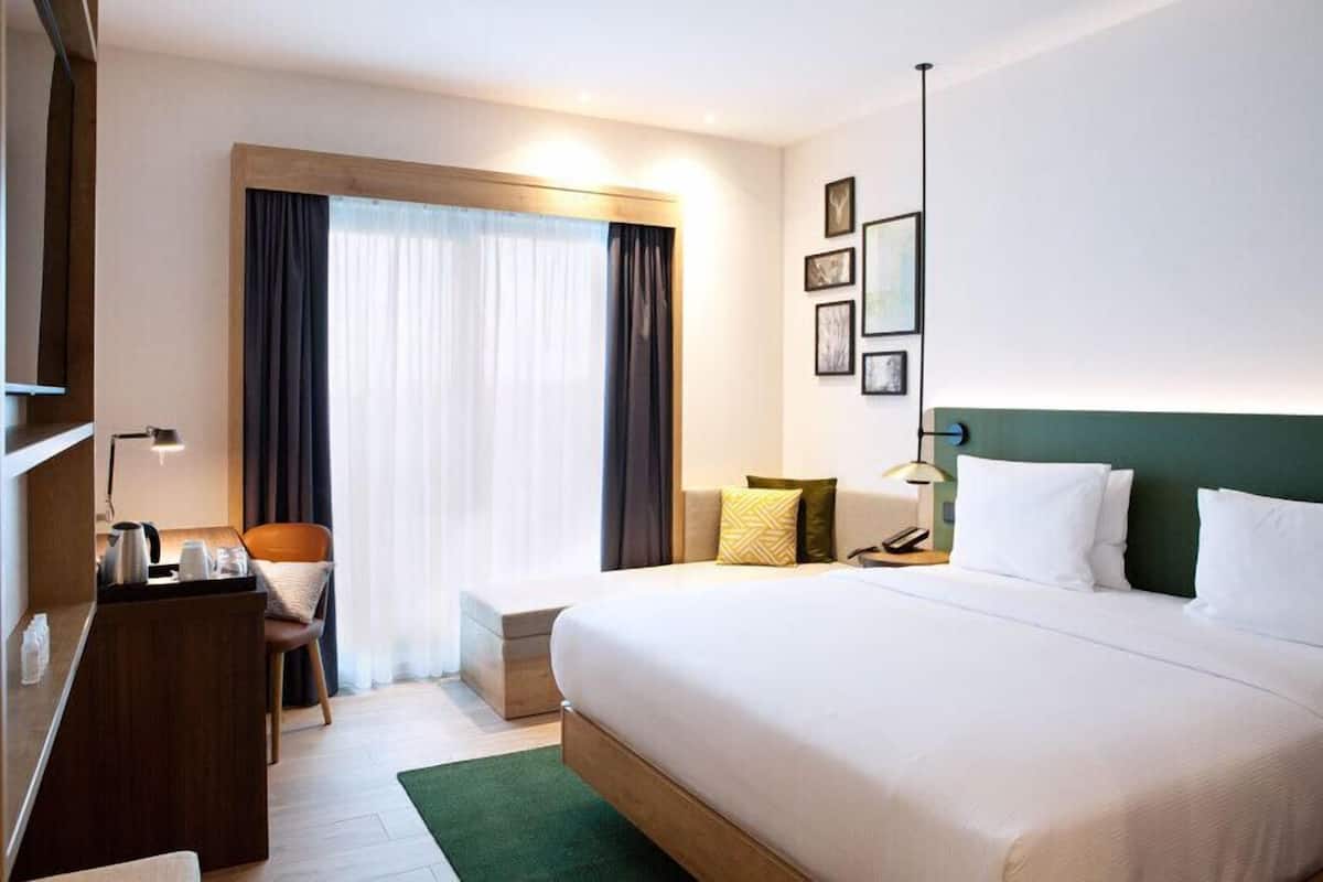 A hotel room with a large bed, a desk with a chair, a small table, and a window with curtains.
