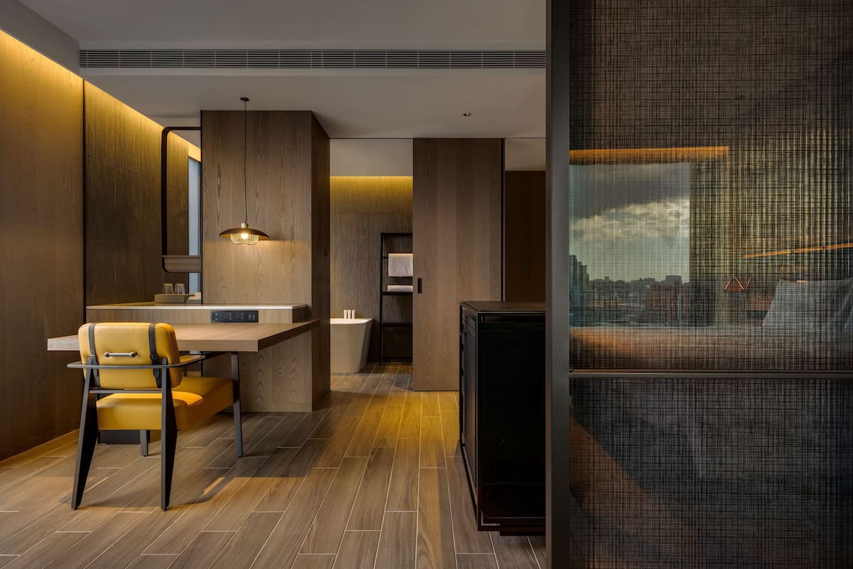 A modern hotel room with a wooden floor, a dining area with a yellow chair, a small kitchen, and a large window with a city view.