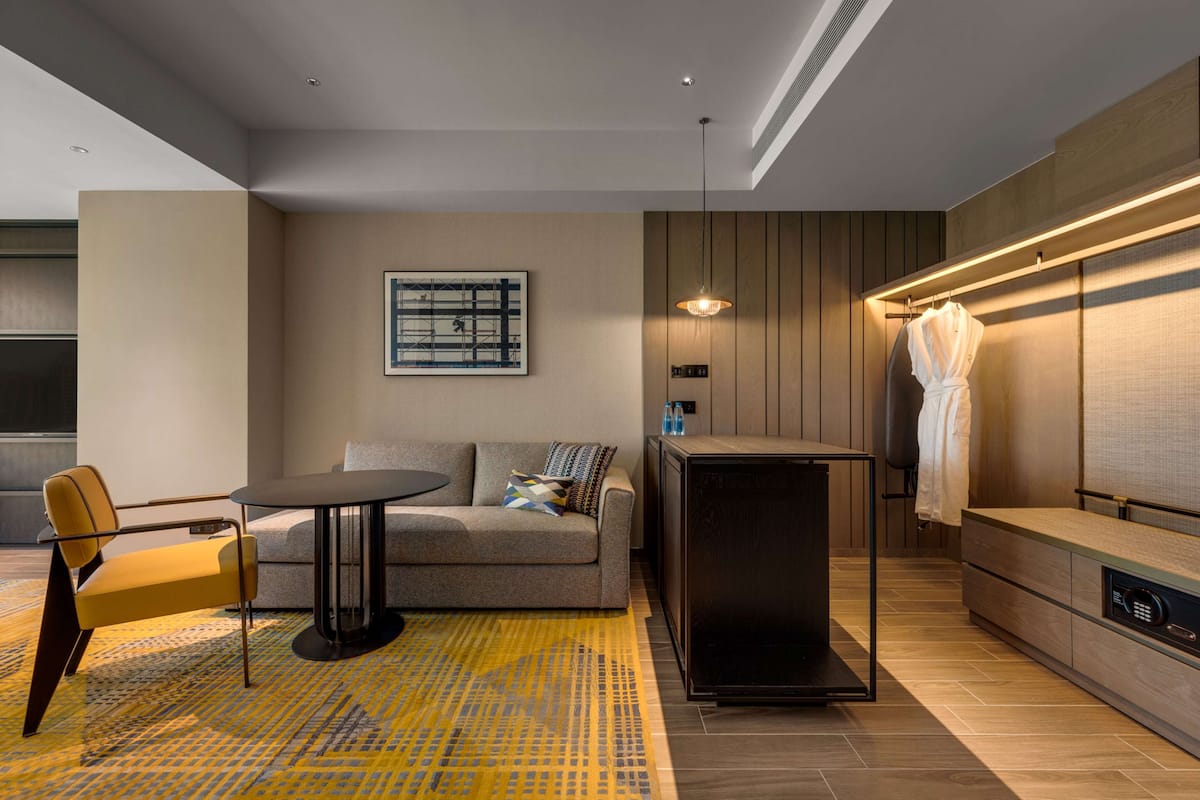 A modern hotel room with a sofa, a small round table, a chair, a built-in heater, and a wall-mounted towel rack.