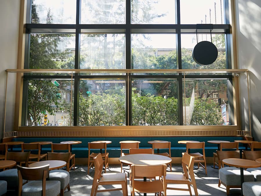 Una cafetería con mesas y sillas de madera, un banco acolchado verde y grandes ventanales con vistas a árboles y edificios.