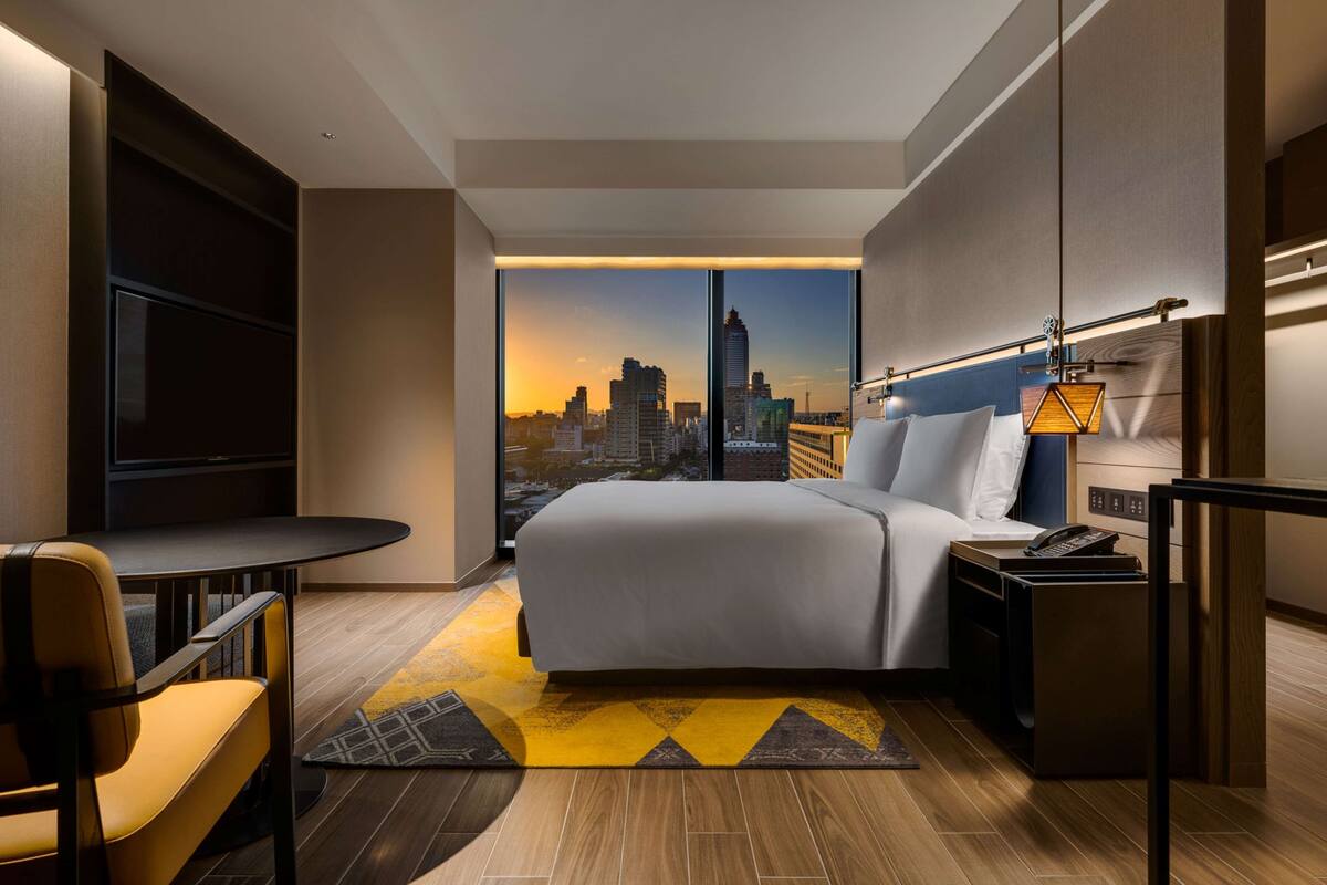 A modern hotel room with a large bed, a desk, a chair, a nightstand, and a view of a city skyline.