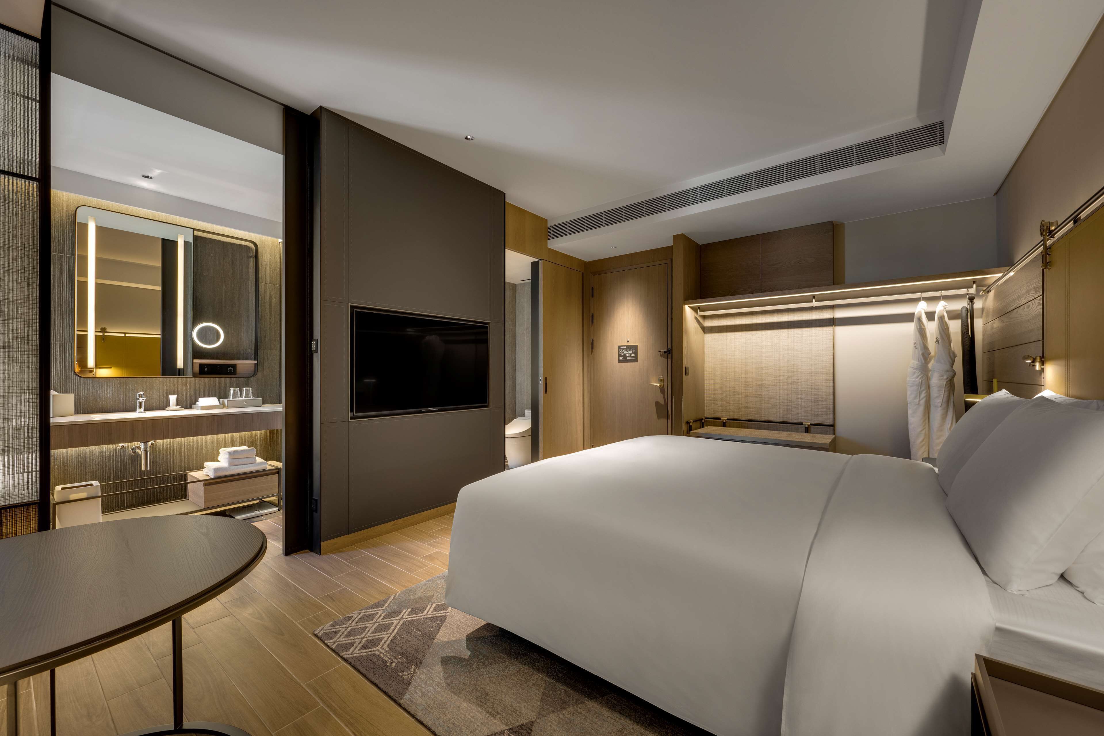 A modern hotel room with a large bed, a TV, a mirror with lights, a bathroom with a toilet, and a small table.