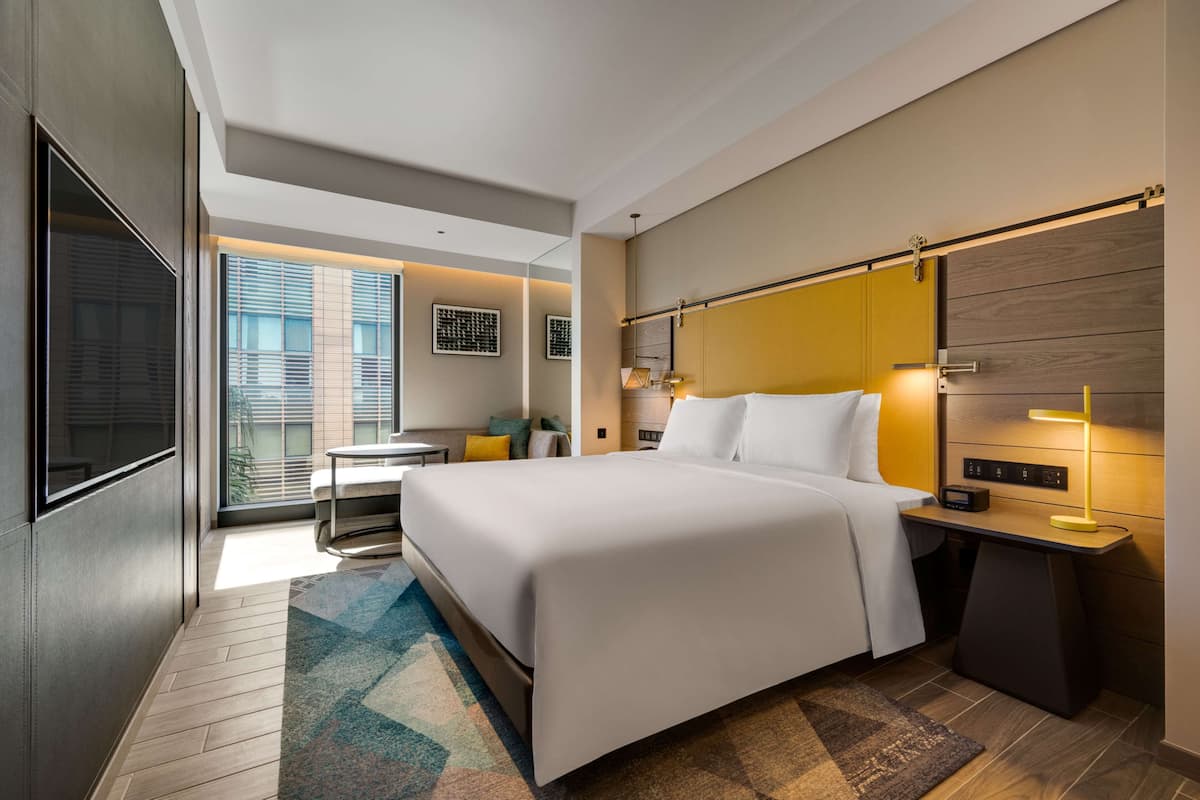 A modern hotel room with a large bed, bedside tables, a desk, and a view of the city through large windows.