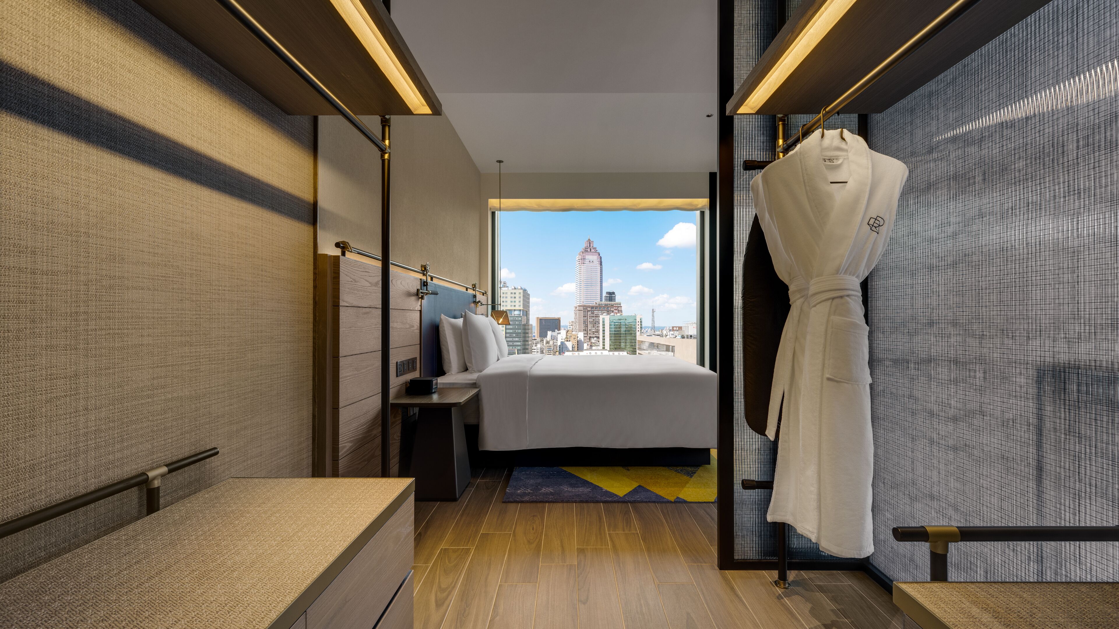 Ein modernes Hotelzimmer mit Blick auf die Stadt, ein Bett, ein Bademantel und ein Badezimmer mit Waschbecken und Spiegel.