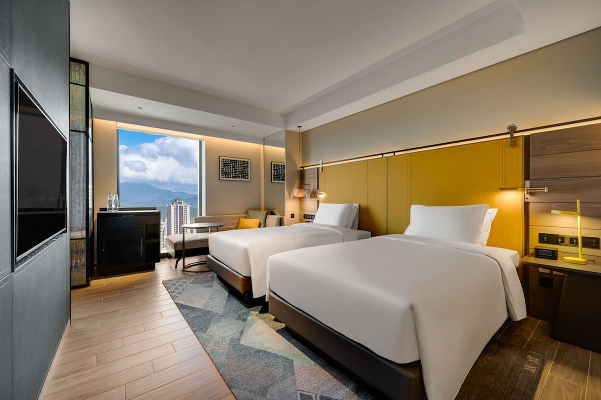 A hotel room with two beds, a desk, a chair, a TV, and a window with a city view.