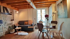 A cozy living room with a wooden ceiling, a stone wall, a sofa, a dining table with chairs, and a painting on the wall. - LA MAISON BALANCOIRE - Private parking - private terrace - La Clé de Honfleur (Honfleur)