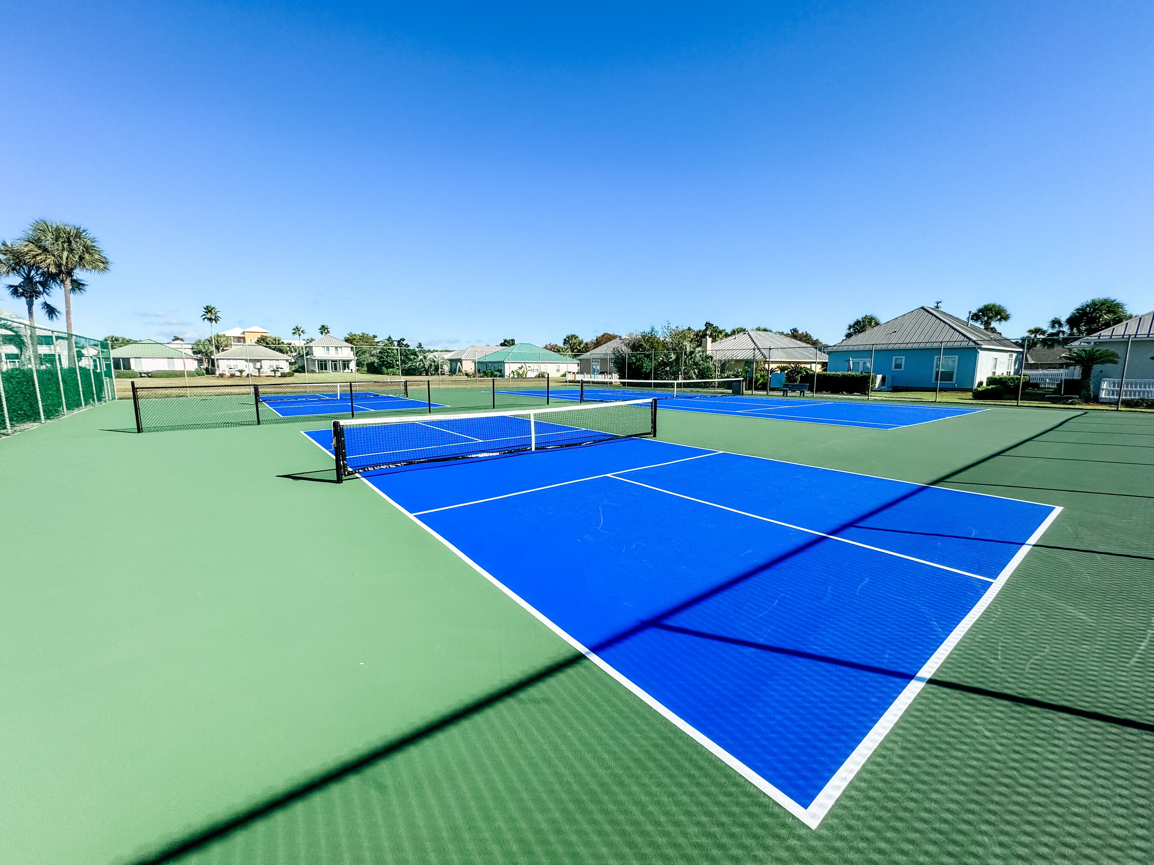 Een tennisbaan met een net, omringd door een hek en huizen op de achtergrond.