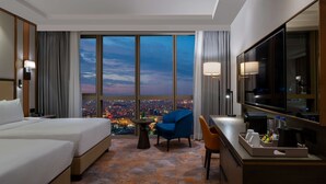 A hotel room with two beds, a desk, a chair, a TV, and a view of a cityscape at dusk.