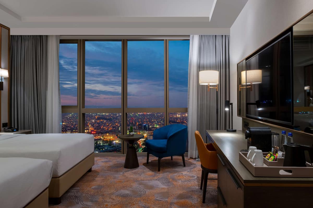 A hotel room with two beds, a desk, a chair, a TV, and a view of a cityscape at dusk.