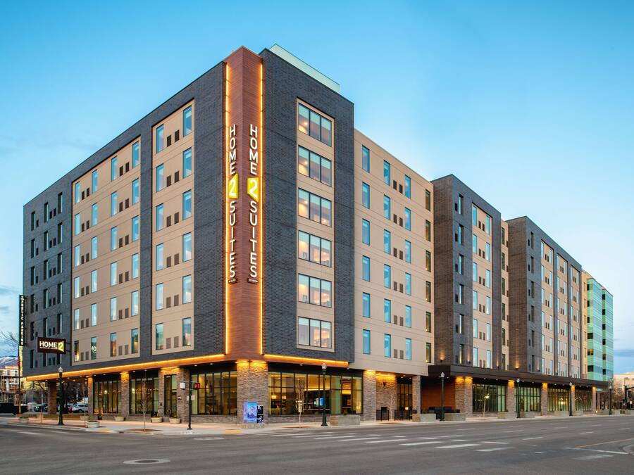 A modern hotel with a prominent 'Home 2 Suites' sign, multiple floors, and a well-lit facade.